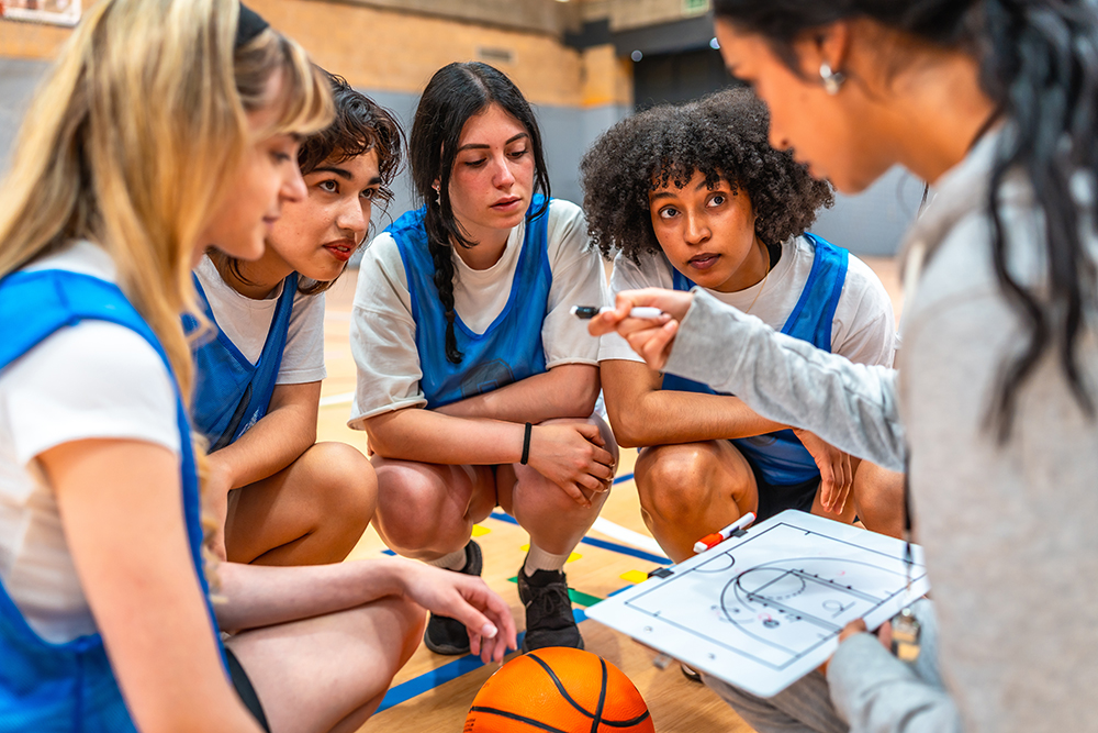 Coach crouches with a group of players in blue practice jerseys, pointing to a whiteboard diagram while explaining a play on an indoor basketball court.